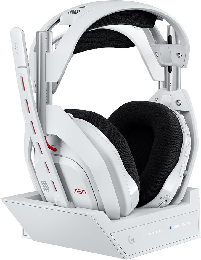 Auriculares gaming blancos Logitech Astro A50 sobre su base de carga inalámbrica junto a mandos de consola